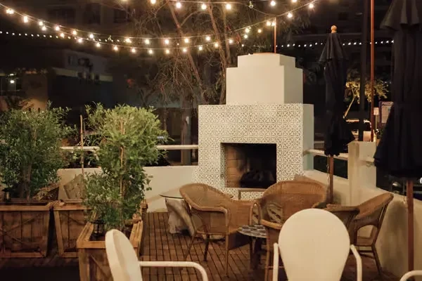 Outdoor night patio with tiled fireplace, wicker chairs, potted plants, lights.