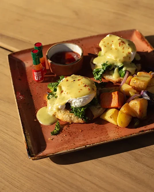 Eggs Benedict with hollandaise, leafy greens, roasted potatoes, hot sauce.