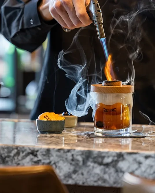 Hand torches cocktail under wooden cap, creating white smoke.