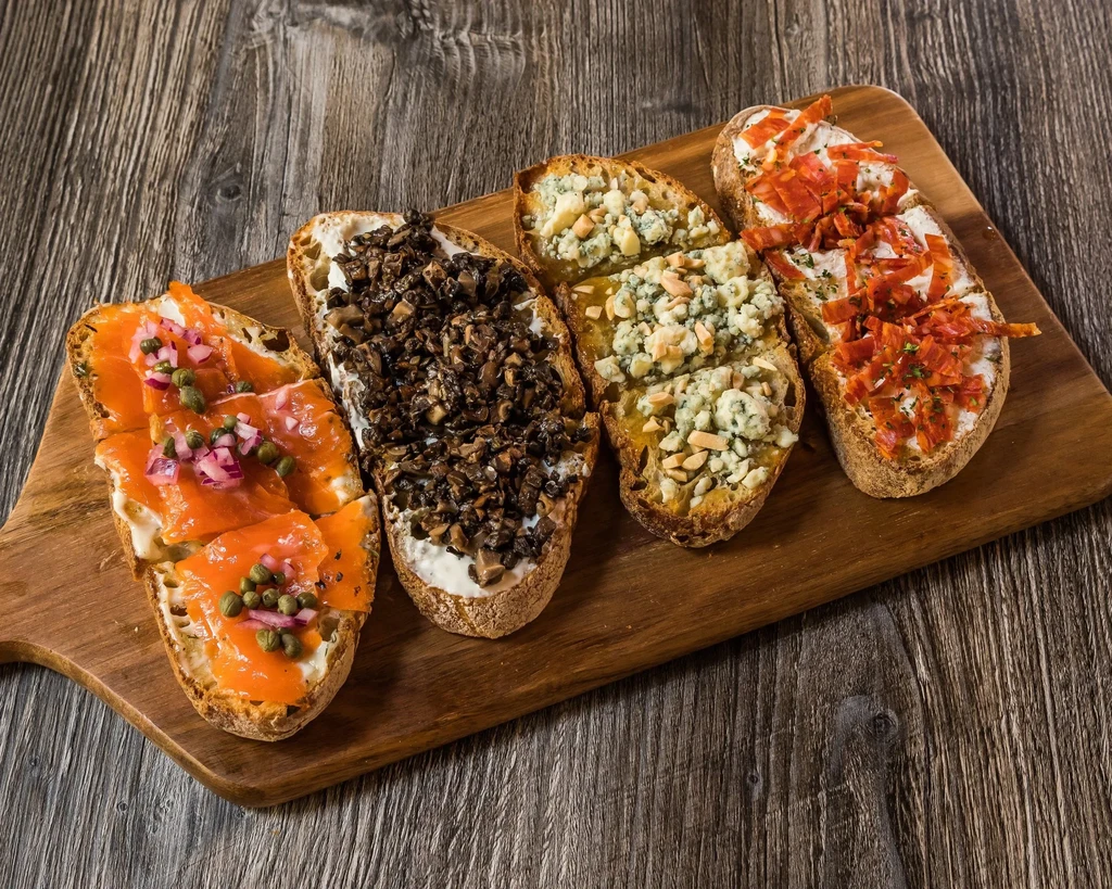 Four bruschetta with diverse toppings on a rustic wooden board.