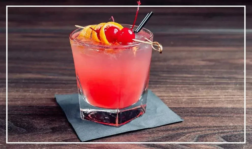Reddish-pink cocktail in rocks glass with cherries, orange peel, and straw.