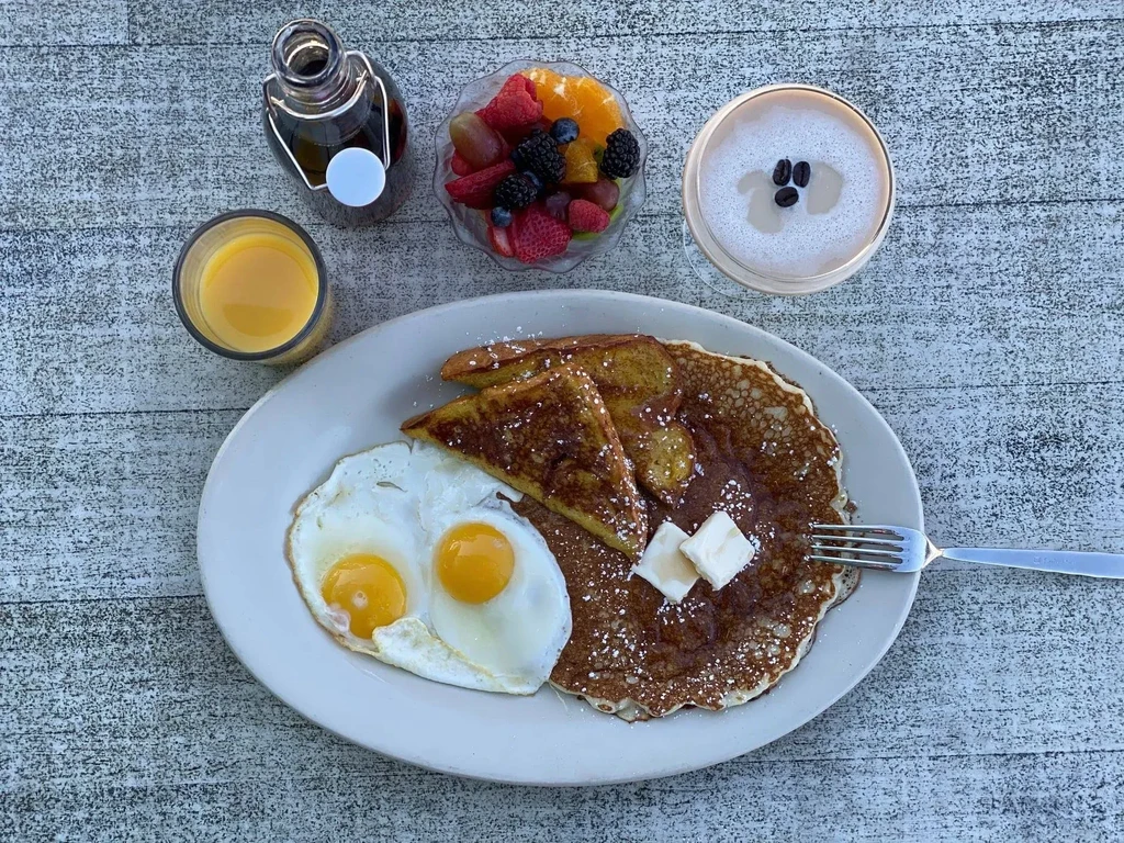 Top-down breakfast spread with eggs, pancakes, French toast, fruit, and drinks.