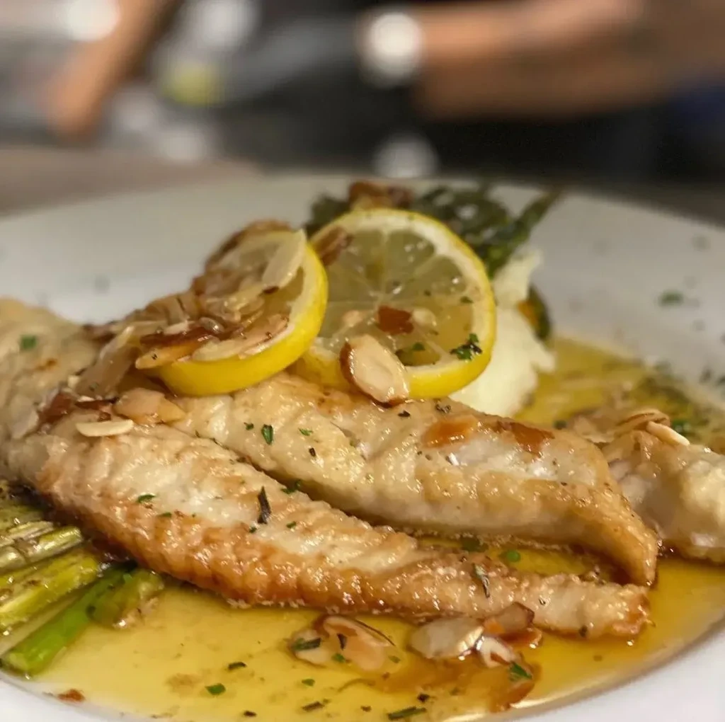 Two golden-brown fish fillets Almandine, lemon, asparagus, and white side.