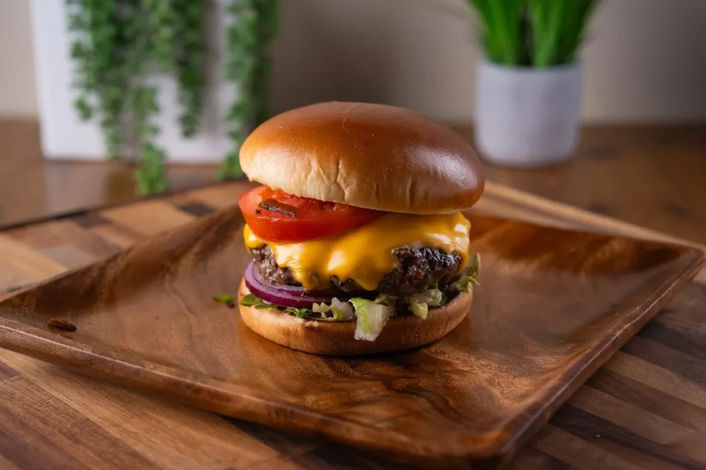 Cheeseburger with brioche bun, melted cheddar, tomato, onion, lettuce on wooden plate.