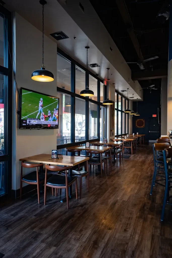 Modern casual restaurant interior with dining tables, windows, and football on TV.