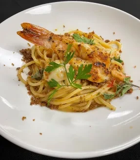 Plated pasta with grilled prawn, smaller seafood, herbs, and crumbs.