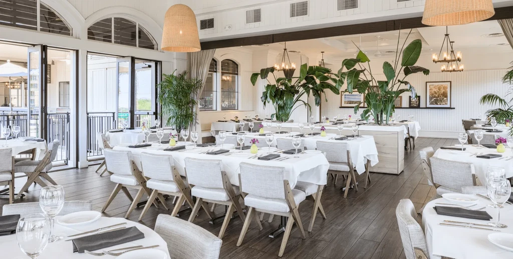 Bright restaurant dining room with set tables, plants, and large windows.