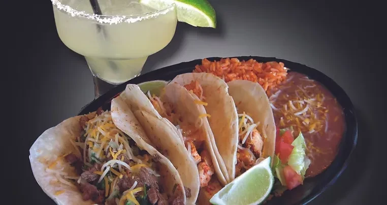 Three tacos, rice, beans, and a margarita on a black plate.