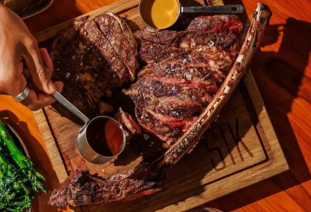 Overhead: Carved medium-rare tomahawk steak, sauces, green vegetables.