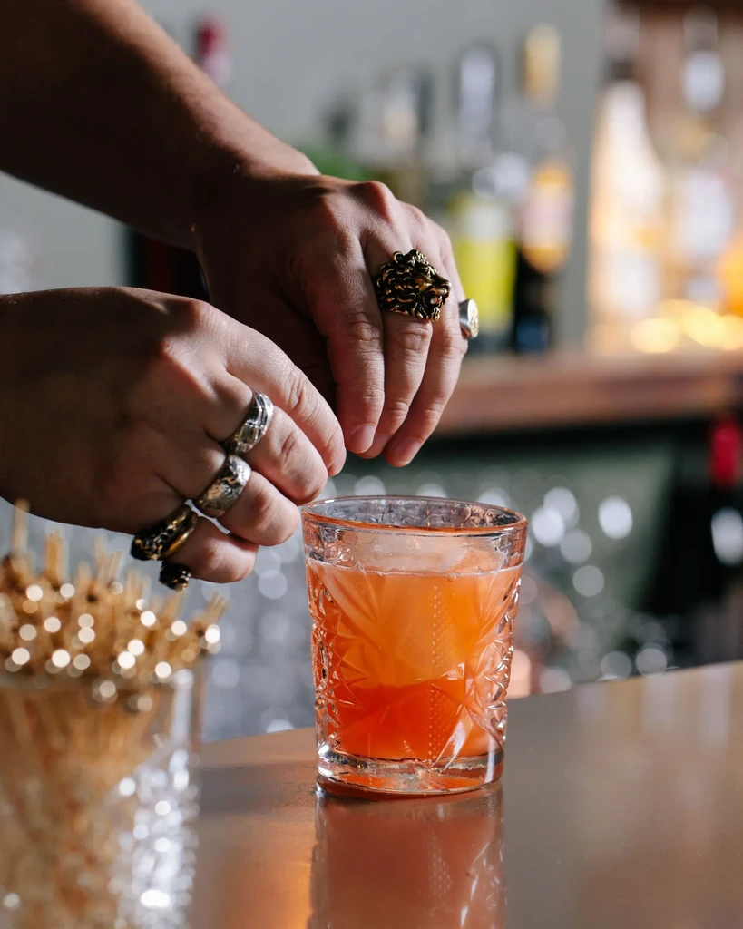 Hands with rings garnish an orange cocktail with ice at a bar.