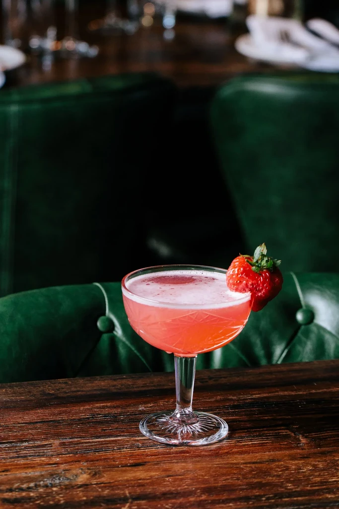Pink cocktail in etched glass with fresh strawberry on wooden table.