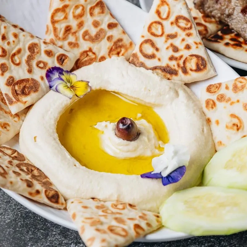 Creamy hummus with olive oil, olive, edible flowers, pita, and cucumber.