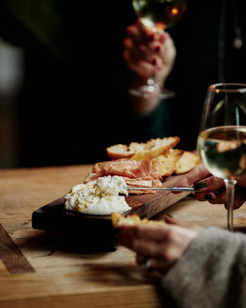 Wooden charcuterie board with cheese, meat, bread; hands reaching, holding wine.