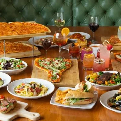 Restaurant table laden with diverse pizzas, pasta, salad, sandwiches, and drinks.