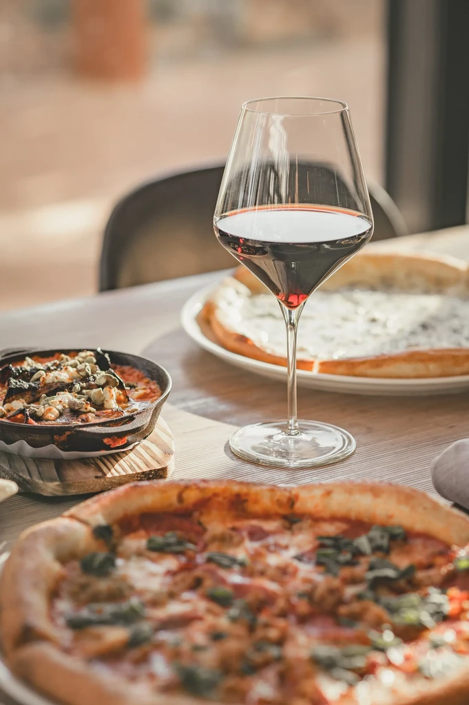 Red wine glass, pizzas, and a savory dish on a dining table.