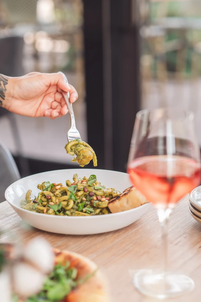 Tattooed hand lifts pesto cavatappi pasta from a bowl with fork.