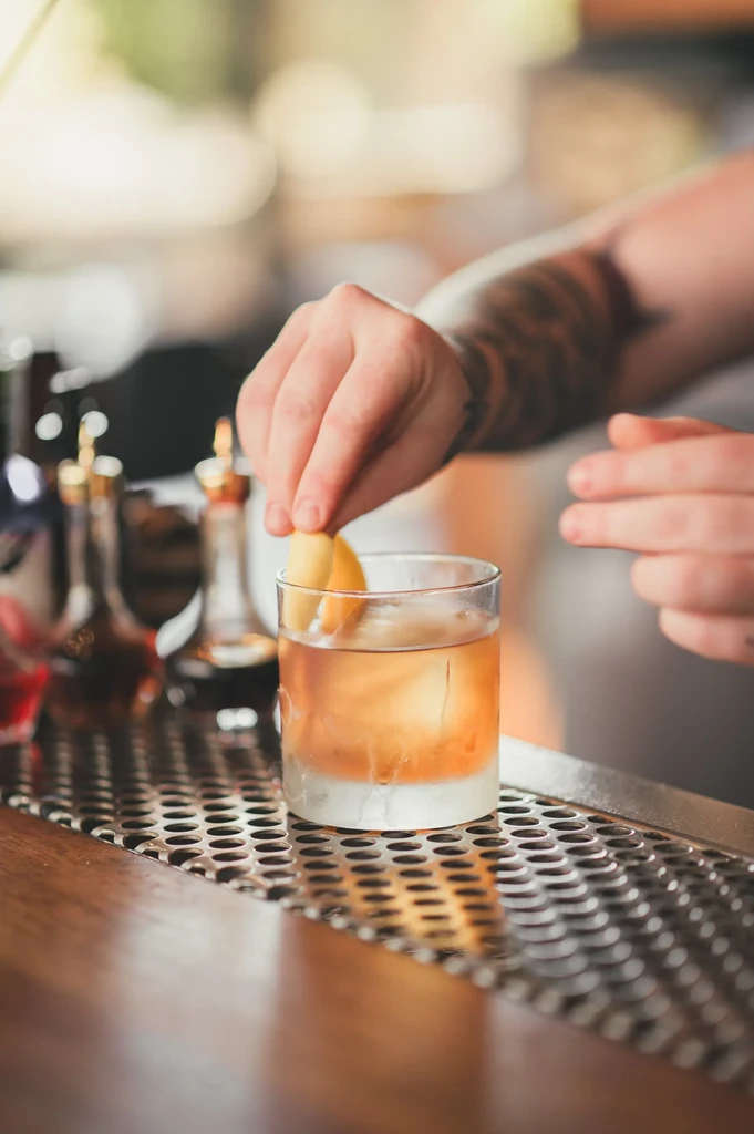 Hands garnish an amber cocktail with a citrus peel at a bar.