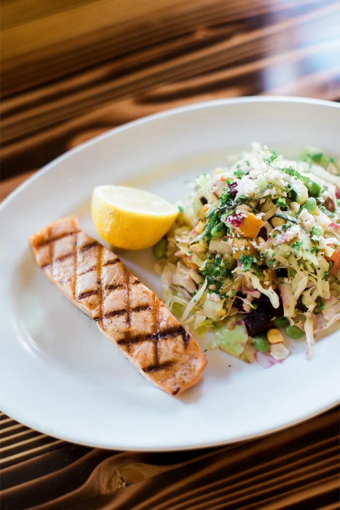 Grilled salmon, vibrant salad with feta and lemon on dark wood.