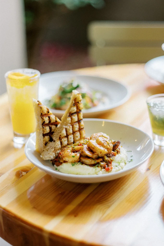 Close-up of shrimp and grits with grilled bread and drinks on a wooden table.