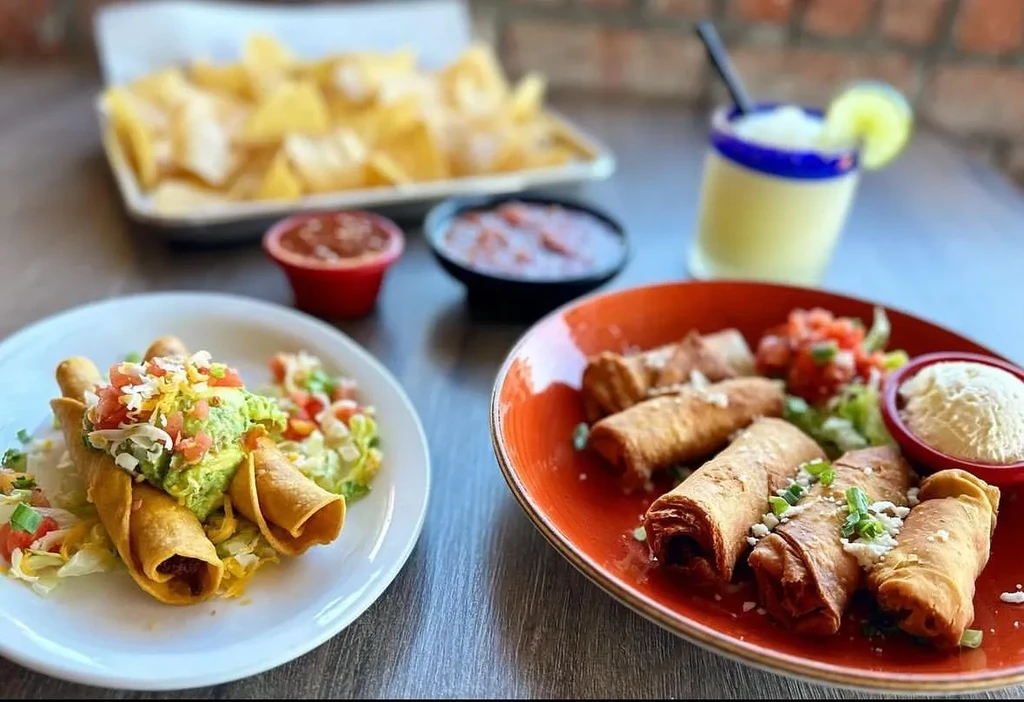 Plates of Mexican taquitos with toppings, chips, salsa, and a drink.