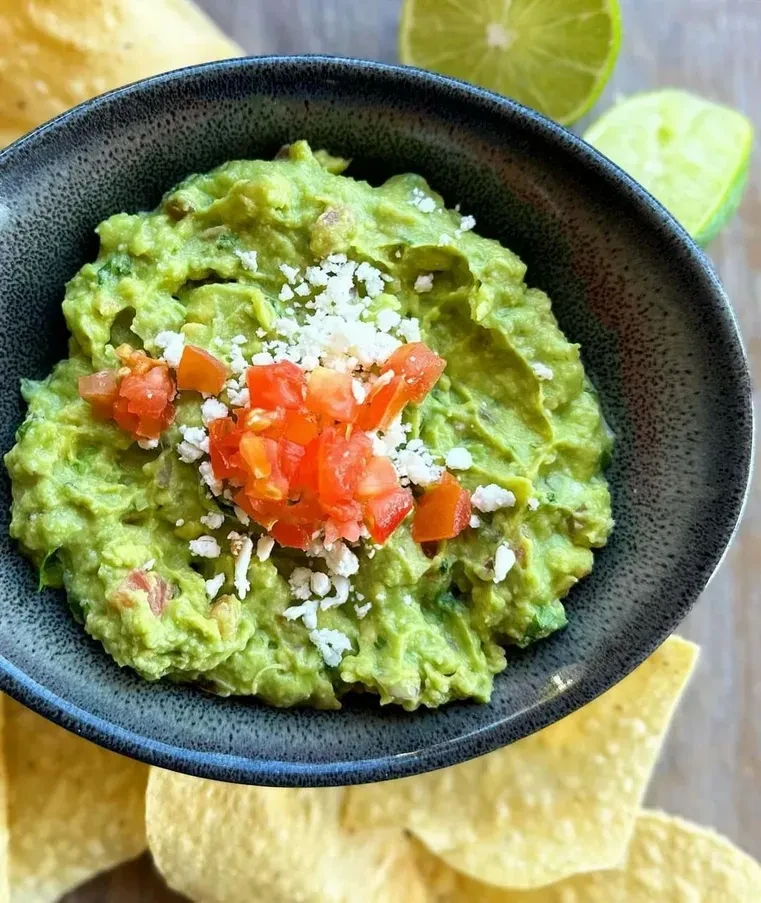 Chunky guacamole with cheese, tomatoes, and limes, surrounded by chips.