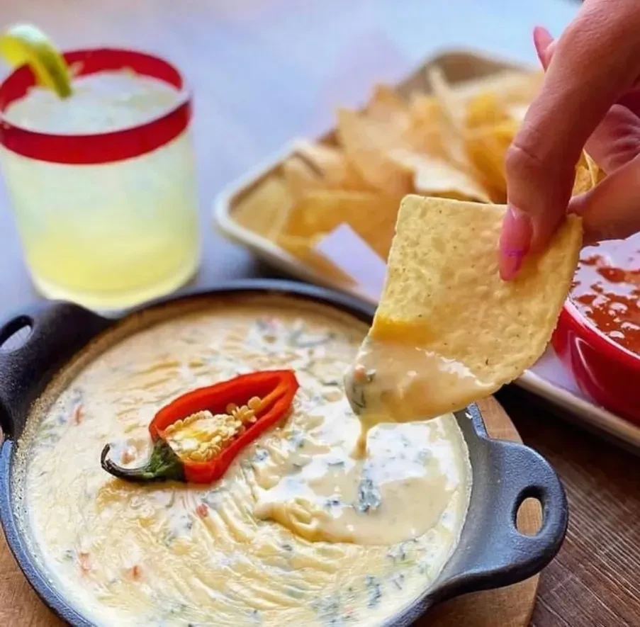 Hand dipping chip into creamy queso skillet, margarita nearby.