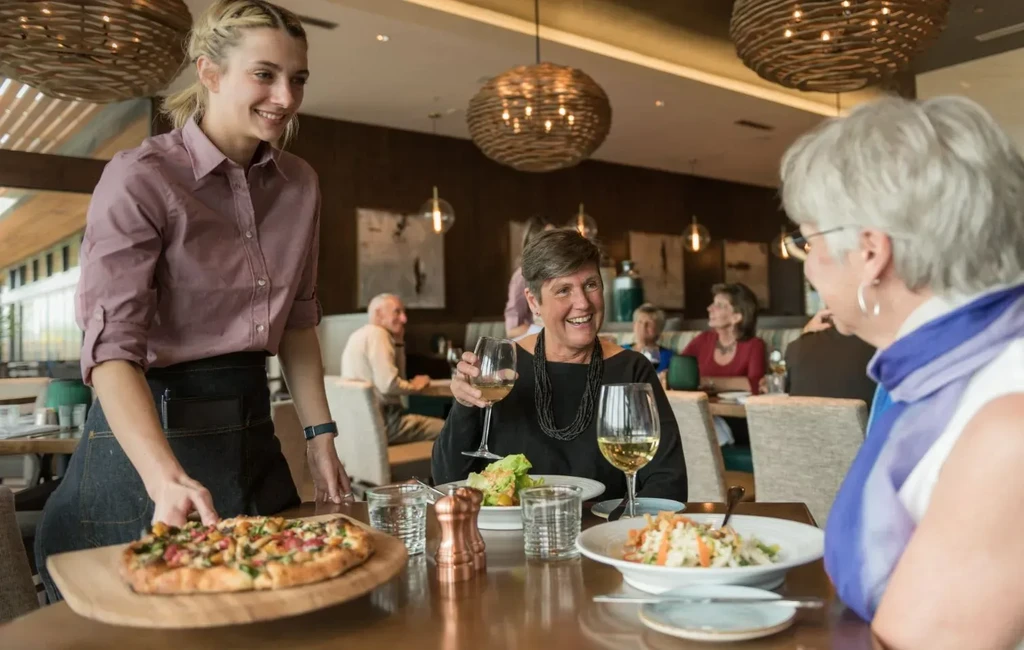 Smiling server presents flatbread pizza to two diners in a restaurant.
