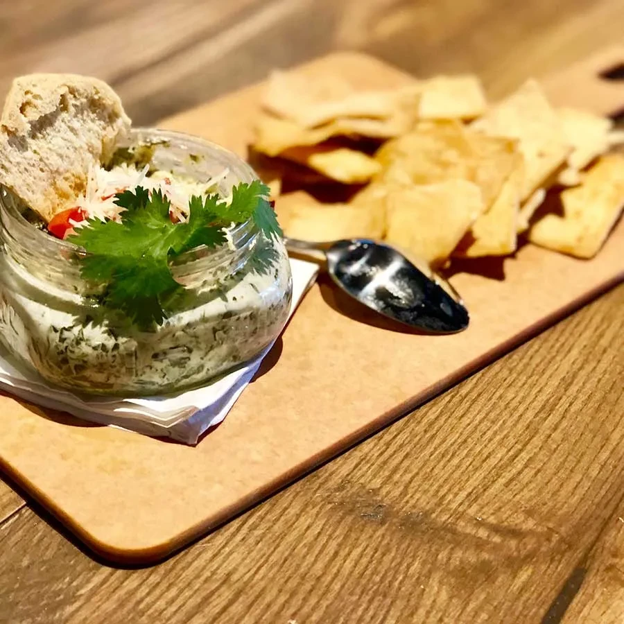 Jar of green dip with bread, cheese, cilantro, and crackers.