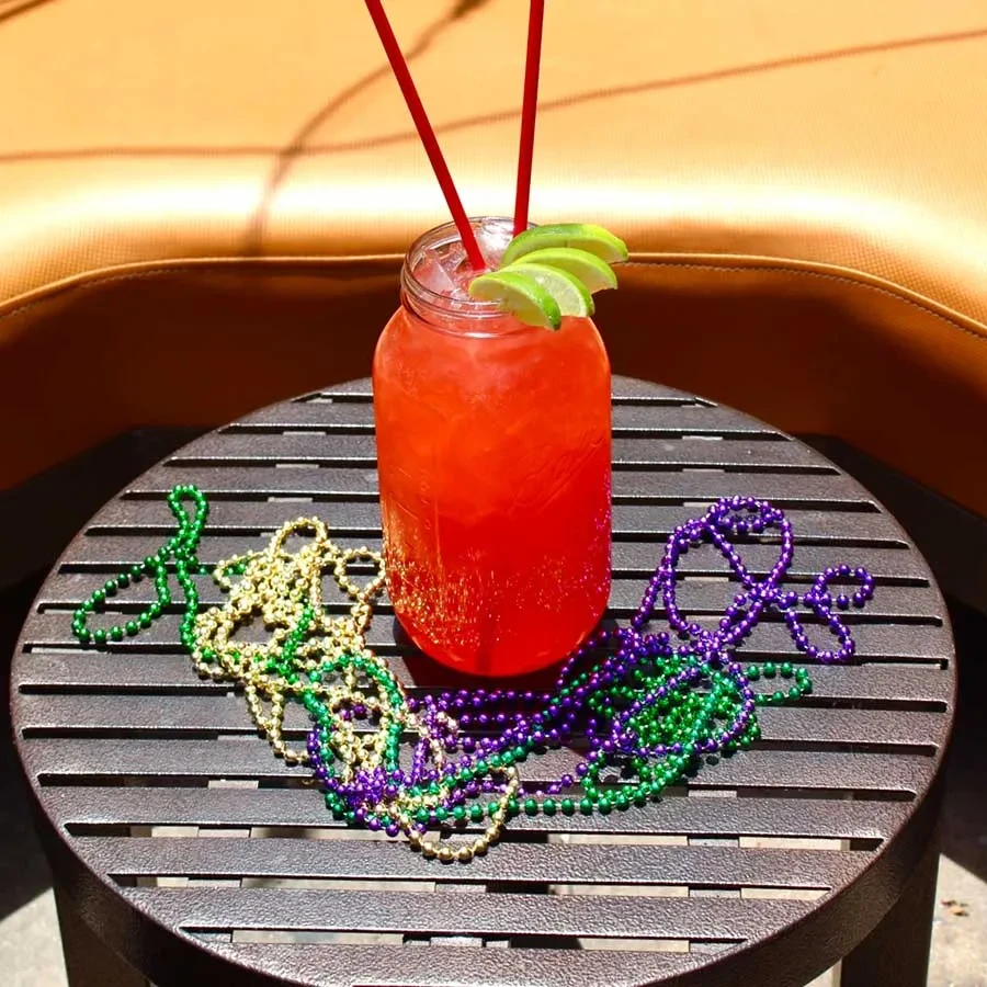 Red-orange cocktail in mason jar with lime and Mardi Gras beads.