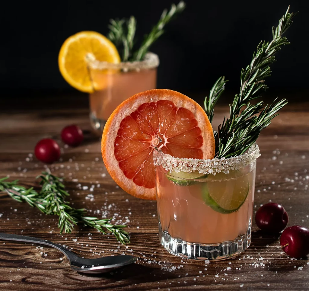 Two cocktails with grapefruit, rosemary, and lime on a rustic wooden surface.
