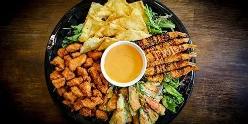 Black platter with varied fried foods, rolls, orange dipping sauce on greens.