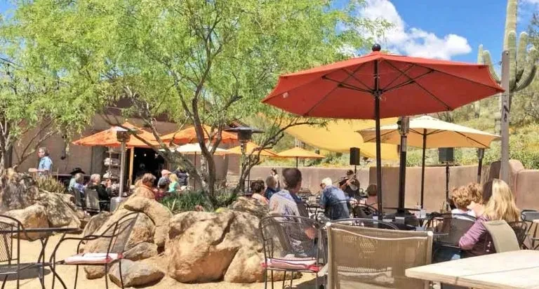 Desert restaurant patio with umbrellas, saguaros, and many people dining.
