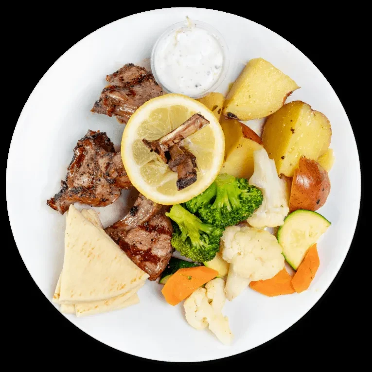 Overhead of grilled lamb chops, roasted vegetables, pita, and white sauce.
