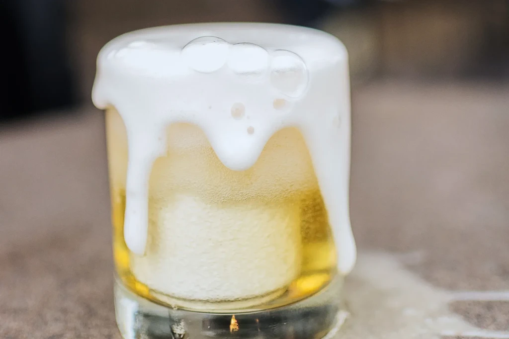 Cold golden beer overflowing with thick white foam from a glass.