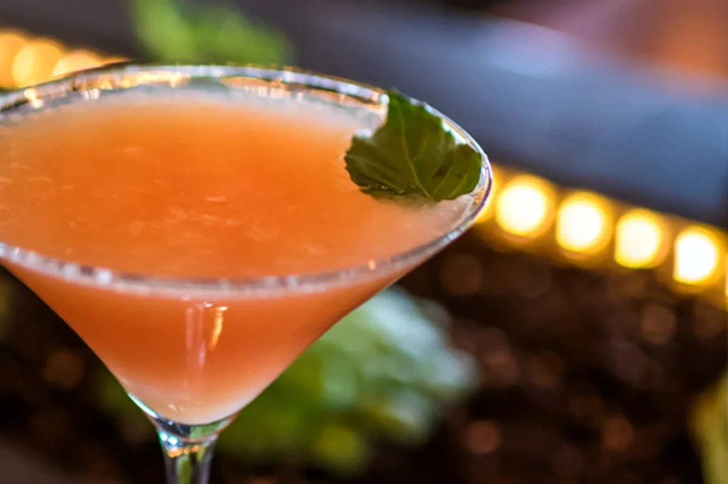 Orange cocktail in martini glass with mint leaf, blurred background.