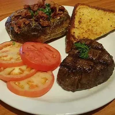 Plate with grilled steak, loaded potato, garlic toast, and tomato slices.