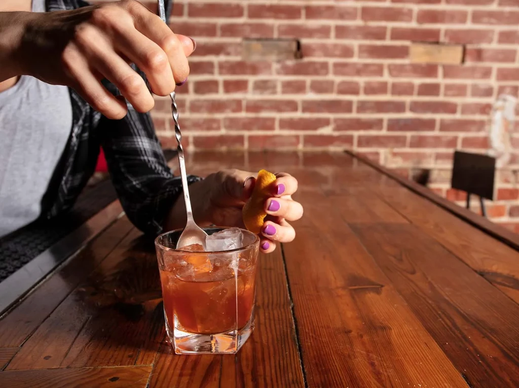 Hands with purple nails stirring an amber drink, garnishing with orange peel.