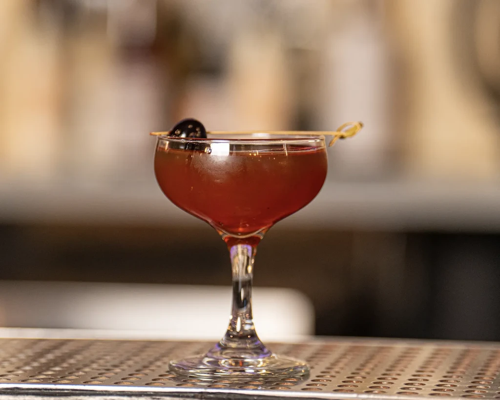 Reddish-brown cocktail in a coupe glass with a cherry garnish.