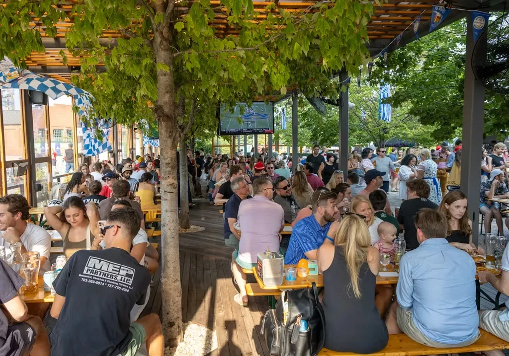 Lively outdoor dining with diverse crowd, Bavarian flags, and sports TV.
