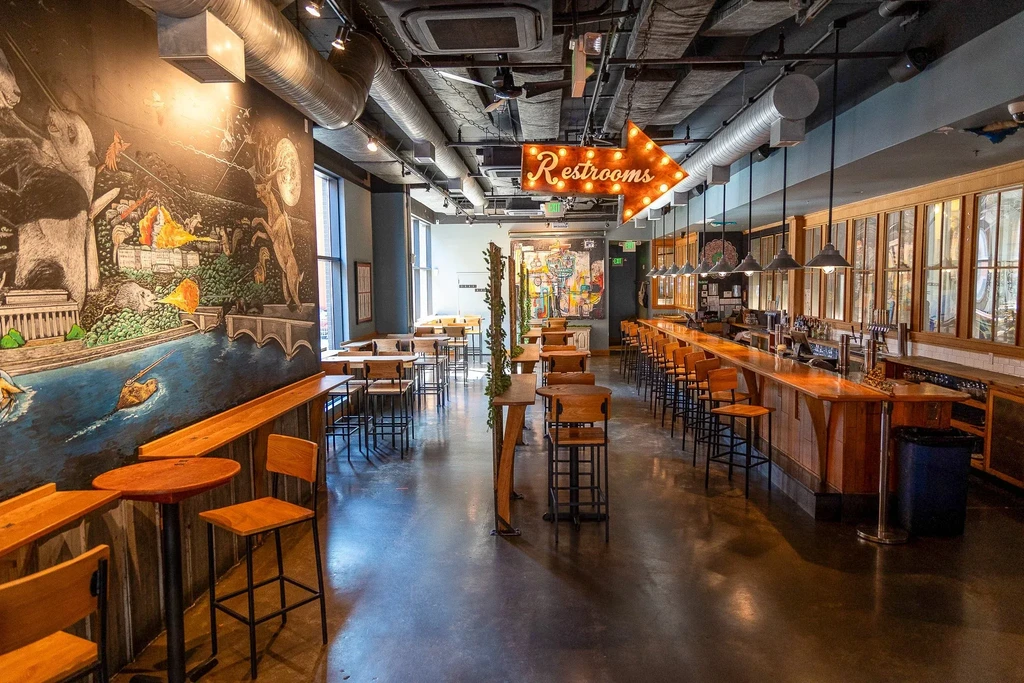 Empty modern restaurant interior with wooden bar, exposed ductwork, and whimsical mural.