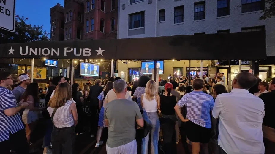 Crowd watches news broadcast on screens outside Union Pub at night.
