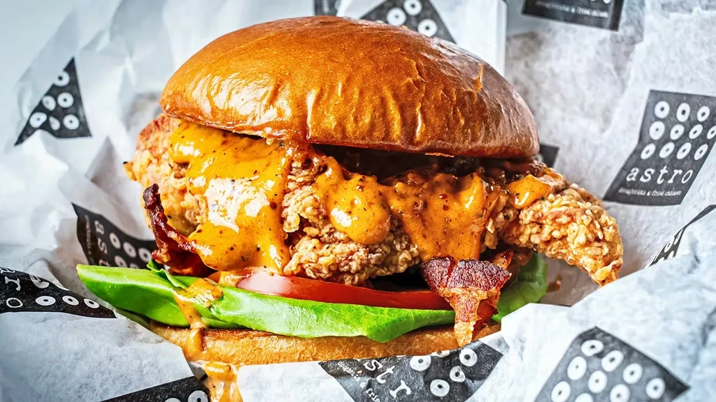 Close-up of a fried chicken sandwich with orange sauce, bacon, lettuce, tomato.