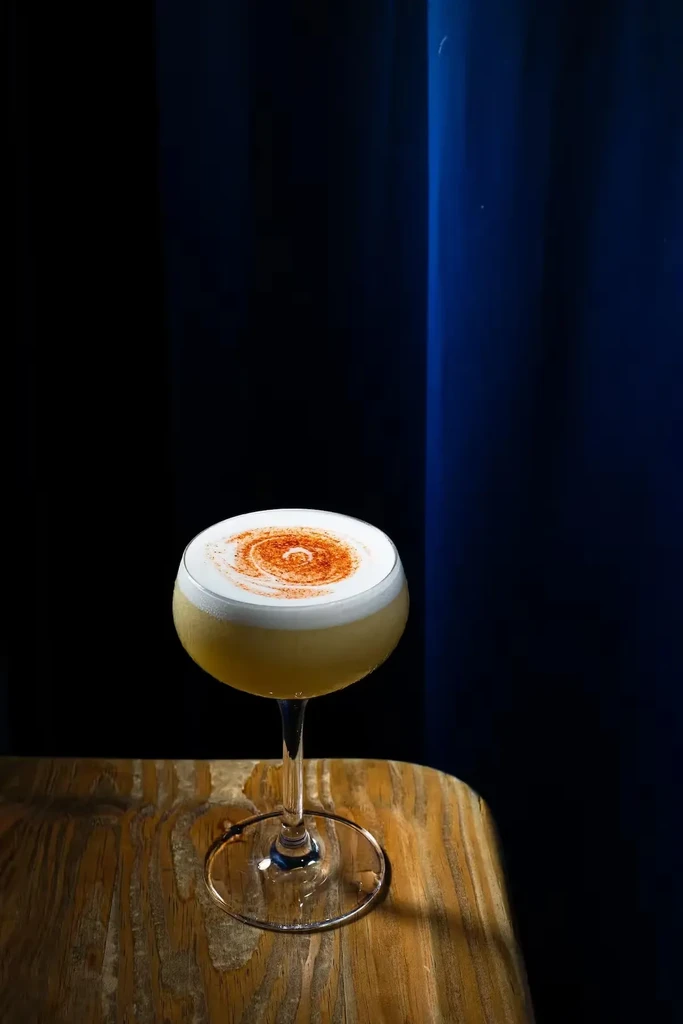 Yellow cocktail with white foam, spiral garnish, on rustic wooden table.