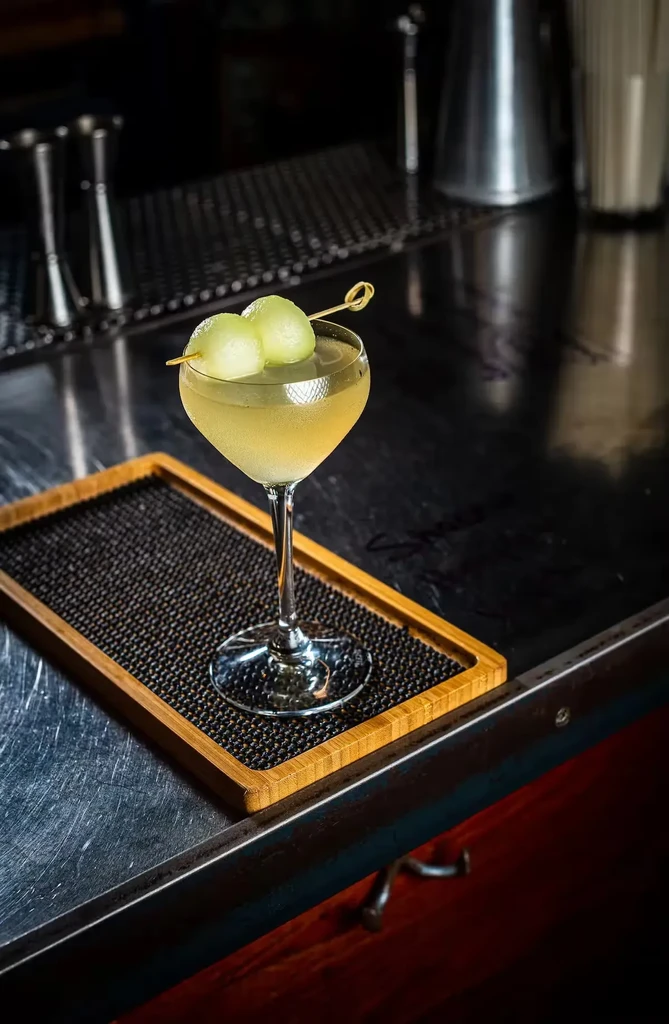 Chilled pale yellow cocktail with melon balls on a bar counter.