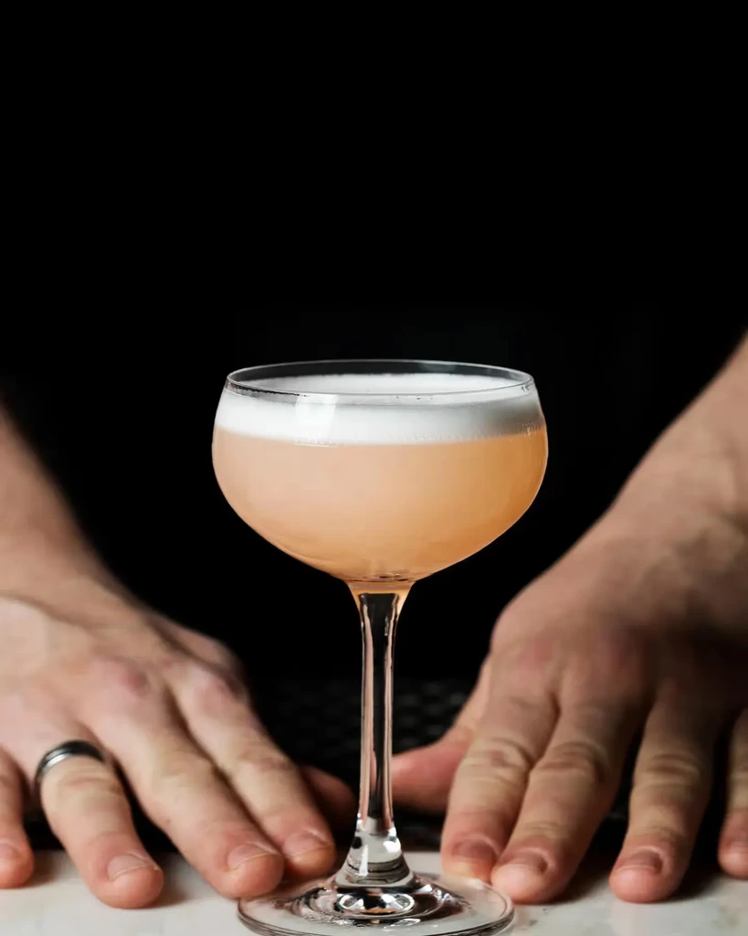 Peach cocktail with white foam in coupe glass, with two hands.