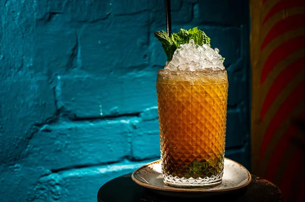 Tall cocktail with crushed ice, mint, and straw on teal brick background.