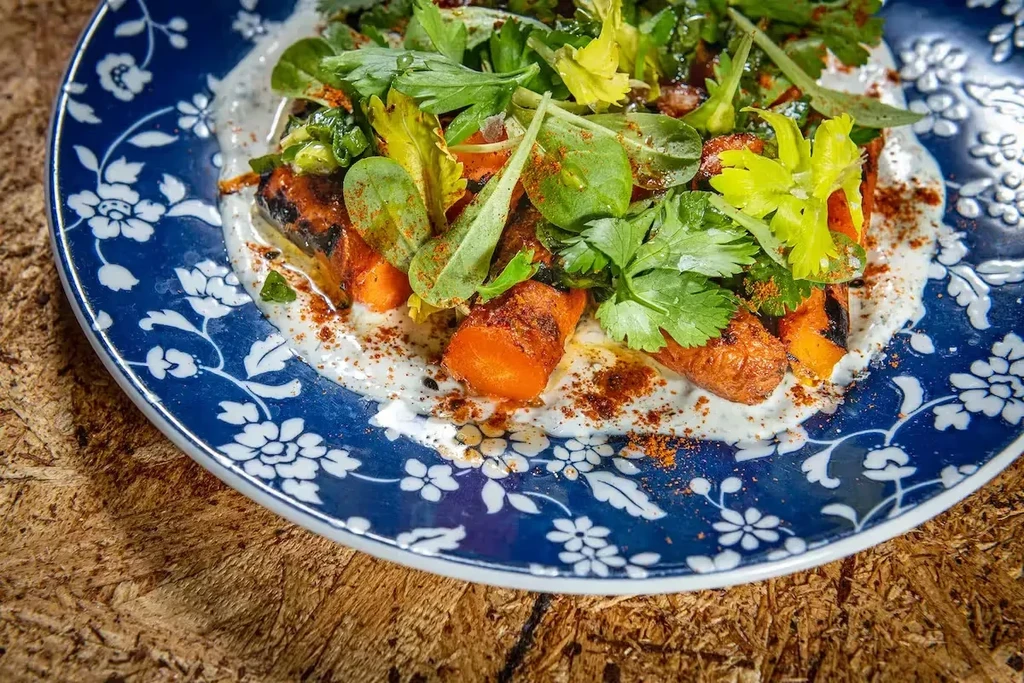 Roasted carrots, creamy sauce, green herbs, and spice on blue plate.