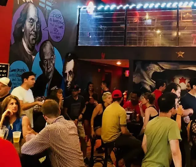 Dark, lively bar scene with patrons, murals, and lights.