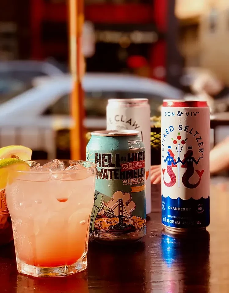 Close-up of a pink cocktail and three seltzer/beer cans on an outdoor table.