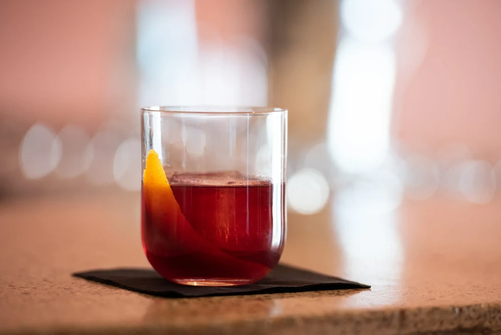 Deep red cocktail in stemless glass, garnished with a citrus peel.